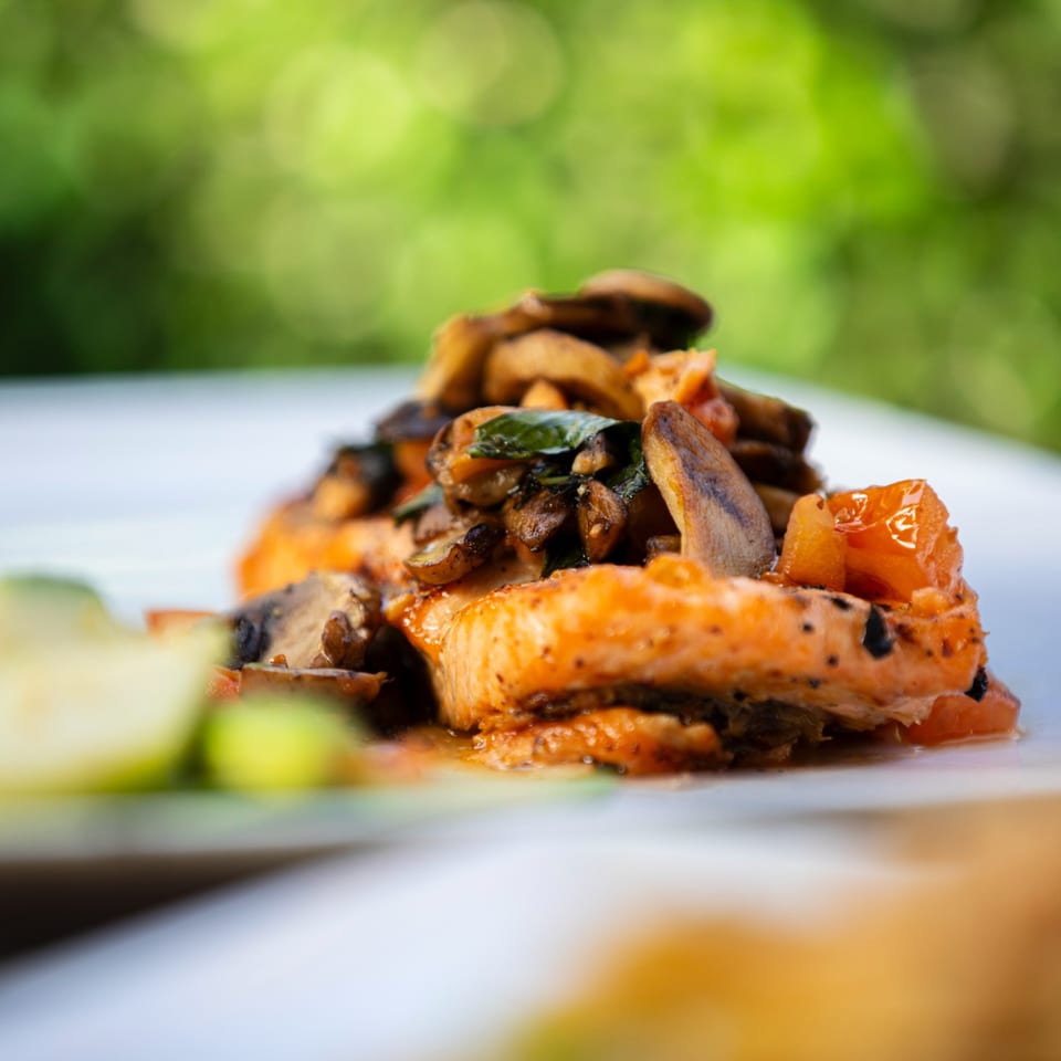 Close-up of salmon fillet topped with sautéed mushrooms and herbs, with a blurred green background.