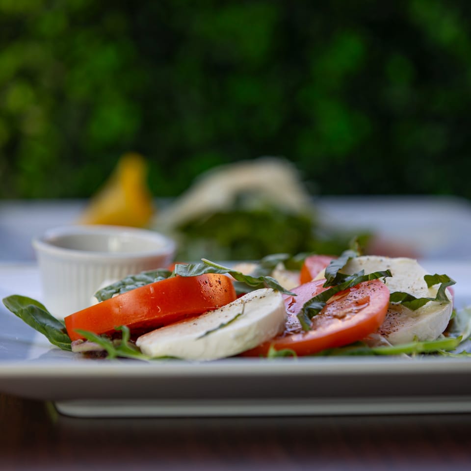 Caprese salad with sliced tomatoes, mozzarella, basil, and a side of dressing on a white plate.