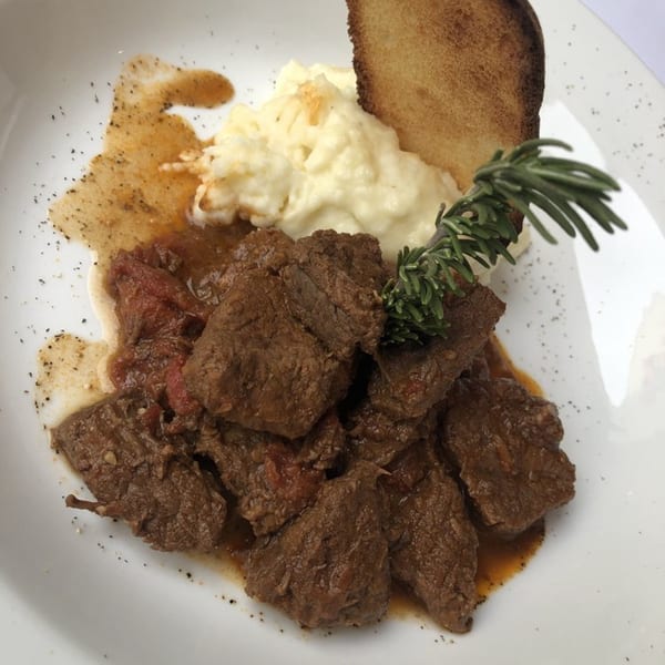 Braised beef chunks with mashed potatoes, a sprig of rosemary, and toasted bread on a white plate.