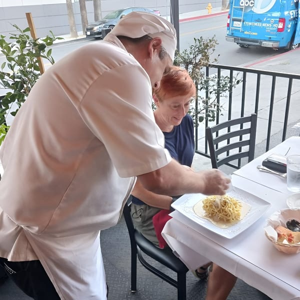 A chef in a white uniform serves a plate of pasta to a smiling woman at an outdoor table.