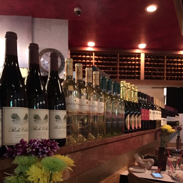 A long row of wine bottles, red and white, on a wooden shelf with flowers and a wine rack in the background.