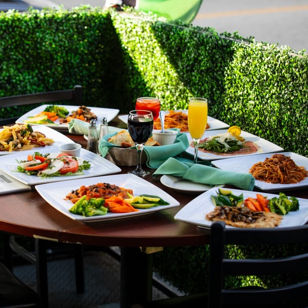 An outdoor table at Pizzeria Vivoli with various Italian dishes, drinks, and bread, surrounded by green hedges.