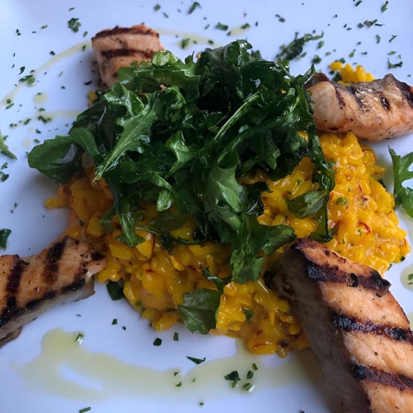 Grilled salmon pieces surround a mound of yellow risotto topped with fresh green arugula.