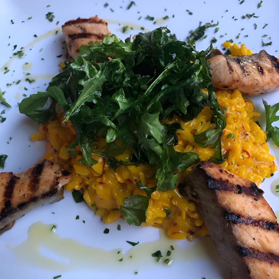 Grilled salmon pieces surround a mound of yellow risotto topped with fresh green arugula.