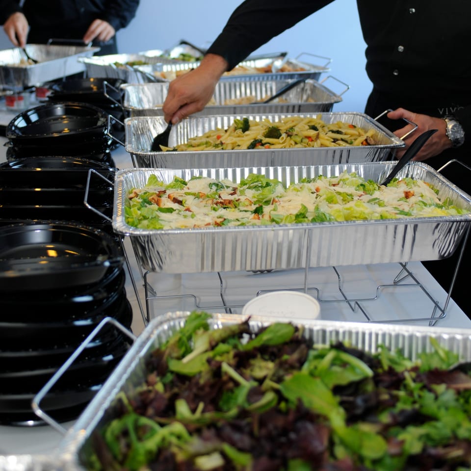 Buffet line with foil pans of pasta, Caesar salad, and mixed greens, with black plates stacked.