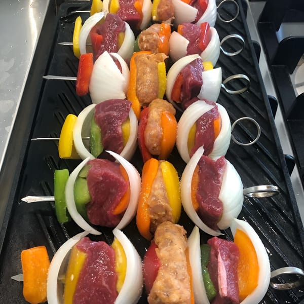 Uncooked beef, sausage, onions, and bell peppers on skewers arranged on a grill.