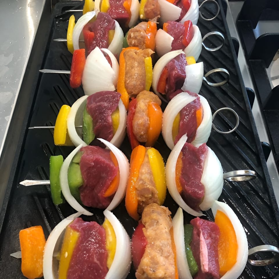 Uncooked beef, sausage, onions, and bell peppers on skewers arranged on a grill.