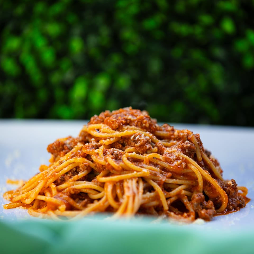Spaghetti bolognese with rich red sauce and grated cheese on a white plate, with a green background.